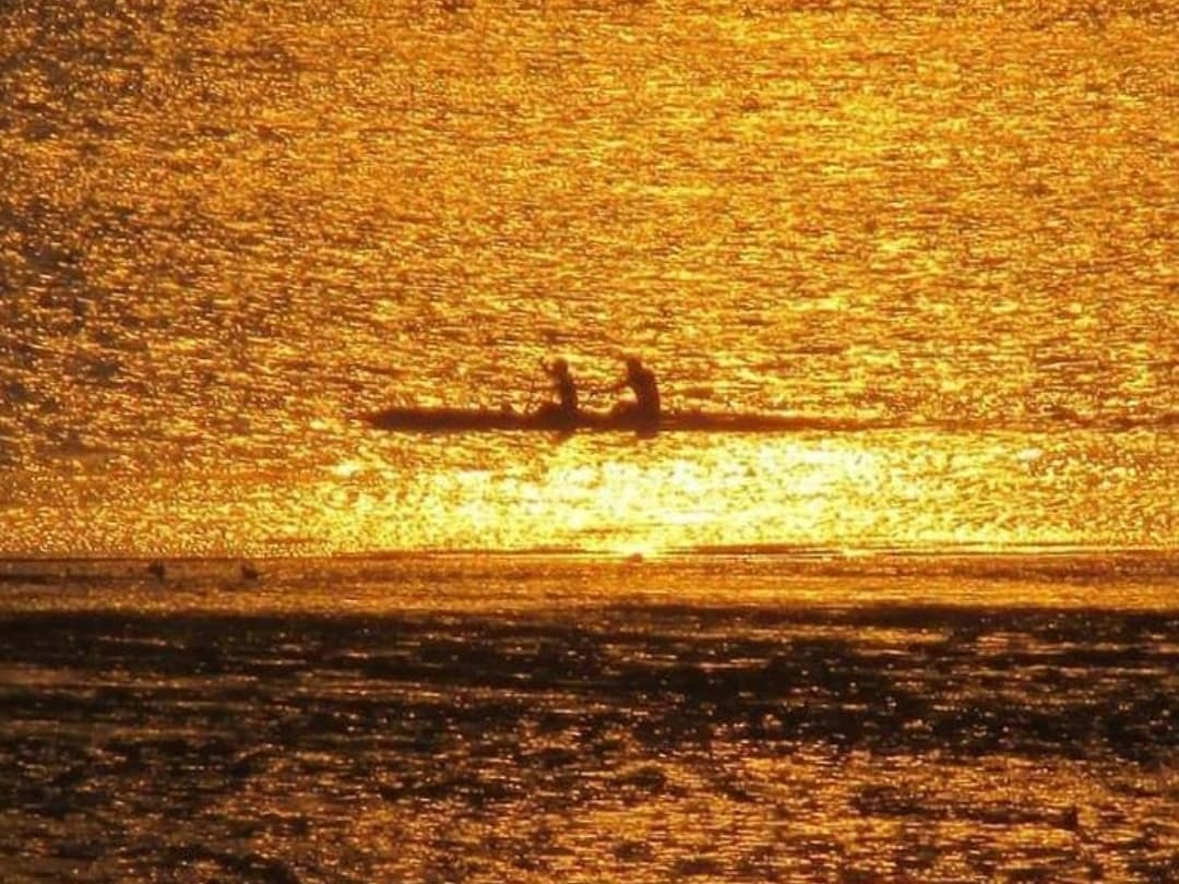 Silhouette eines Outrigger-Kanus mit zwei Personen bei tiefstehender Sonne, die das Wasser in goldenem Licht glitzern lässt.