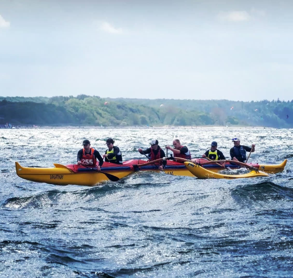 Sechs Paddler in einem gelben Outrigger-Kanu „Vanatinai“ bei Wellengang vor einer bewaldeten Küstenlinie.