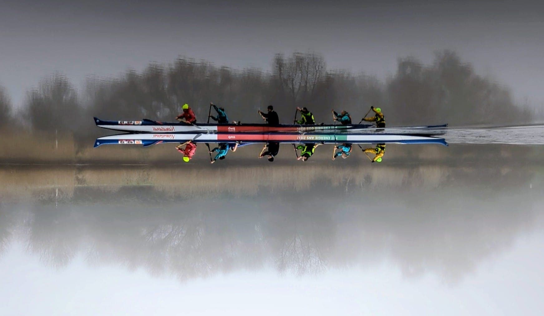 Ein Outrigger-Kanu mit sechs Paddlern auf spiegelglattem Wasser im Nebel, perfekt symmetrische Spiegelung an der Wasseroberfläche.